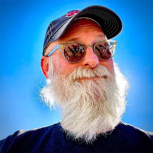 Headshot of Andrew. He is backlit against a brilliant blue sky, sun shining through his long white beard. He’s wearing a Jeep ballcap and sunglasses and has a quizzical smirk on his face.